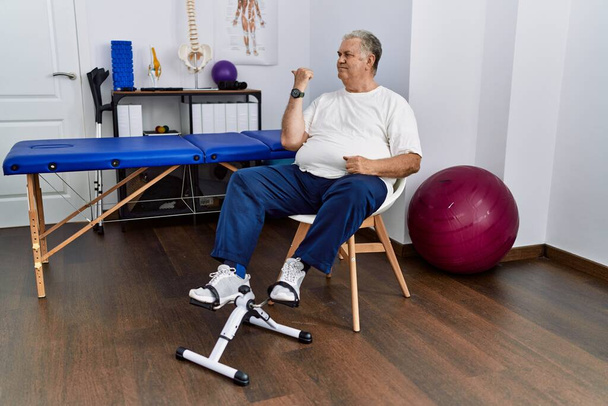 Старший кавказький чоловік у фізіотерапевтичній клініці використовує педальну вправу, посміхаючись із щасливим обличчям, дивлячись і вказуючи на бік великим пальцем вгору
.  - Фото, зображення
