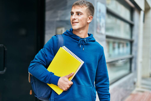 Mladý blond student usměvavý šťastný držení knihy a notebook na univerzitě. - Fotografie, Obrázek