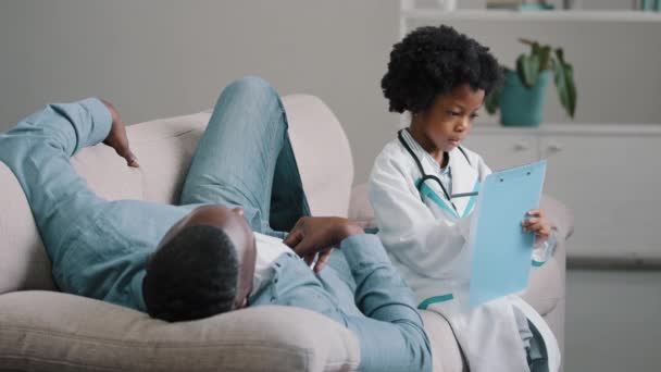 Bambino serio finge di essere medico in costume medico seduto a scrivere sintomi sul tablet padre si trova sul divano si lamenta di sentirsi male sensazioni dolorose papà afro-americano che gioca con la figlia - Filmati, video