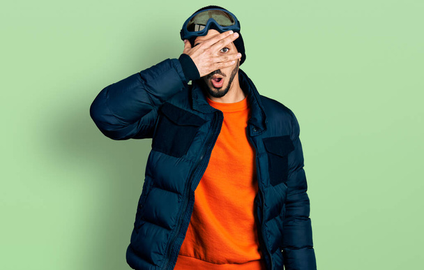 Jeune homme hispanique barbu portant des vêtements de neige et des lunettes de ciel regardant dans le choc couvrant le visage et les yeux avec la main, regardant à travers les doigts avec une expression embarrassée.  - Photo, image
