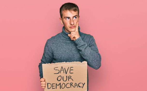 Young blond man holding save our democracy protest banner serious face thinking about question with hand on chin, thoughtful about confusing idea  - 写真・画像