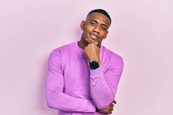 Молодий чорний чоловік у повсякденному рожевому светрі впевнено дивиться на камеру, посміхаючись схрещеними руками та рукою, піднятою на підборідді. мислення позитивне
.  - Фото, зображення