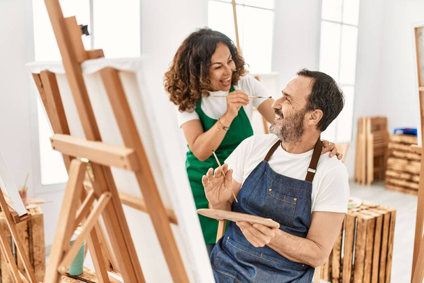 Δύο μαθητές μέσης ηλικίας χαμογελώντας ευτυχισμένοι παίζοντας με ζωγραφική πινέλο ο ένας στον άλλο στη Σχολή Καλών Τεχνών. - Φωτογραφία, εικόνα