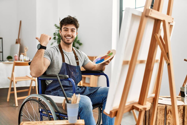 Jeune homme hispanique assis sur la peinture en fauteuil roulant au studio d'art souriant avec le visage heureux regardant et pointant vers le côté avec le pouce vers le haut.  - Photo, image