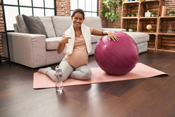 Молода вагітна жінка сидить на йога килимок з пілатесом м'яч вказує пальцем на одного себе посміхаючись щасливим і гордим
  - Фото, зображення