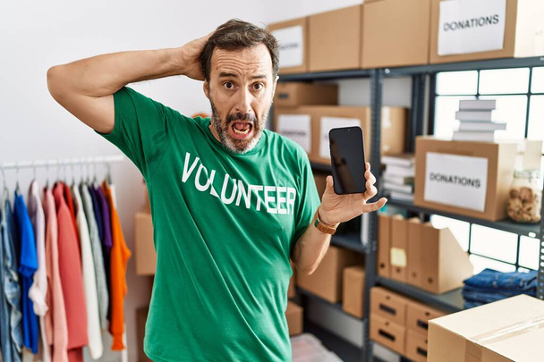 Middle age man with beard wearing volunteer t shirt holding smartphone crazy and scared with hands on head, afraid and surprised of shock with open mouth  - Foto, Bild
