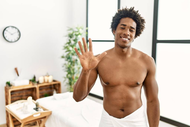 Młody, afrykański mężczyzna bez koszulki, noszący ręcznik w centrum piękności, pokazujący i wskazujący palcami numer pięć, uśmiechając się z pewnością siebie i szczęściem..  - Zdjęcie, obraz