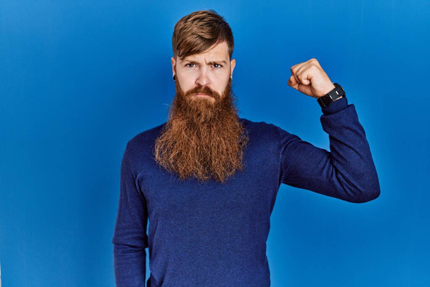 Redhead man with long beard wearing casual blue sweater over blue background strong person showing arm muscle, confident and proud of power  - Valokuva, kuva