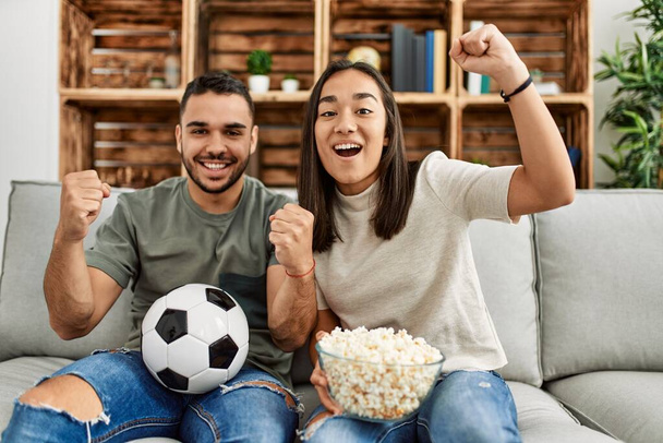 Joven pareja latina viendo un partido de fútbol comiendo palomitas en casa. - Foto, Imagen