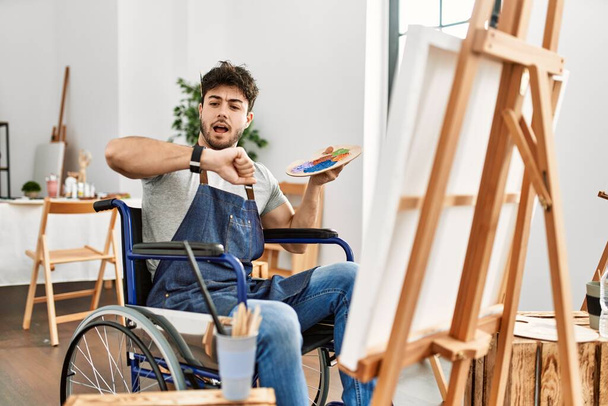 Joven hombre hispano sentado en la pintura en silla de ruedas en el estudio de arte mirando el tiempo del reloj preocupado, con miedo de llegar tarde  - Foto, imagen