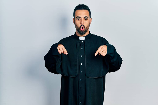 Handsome hispanic man with beard wearing catholic priest robe pointing down with fingers showing advertisement, surprised face and open mouth  - Φωτογραφία, εικόνα