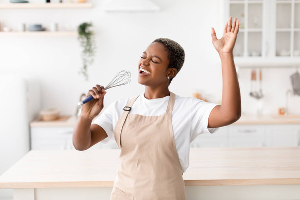 Happy inspired young african american woman in apron prepares food, sings at imaginary microphone - Фото, зображення