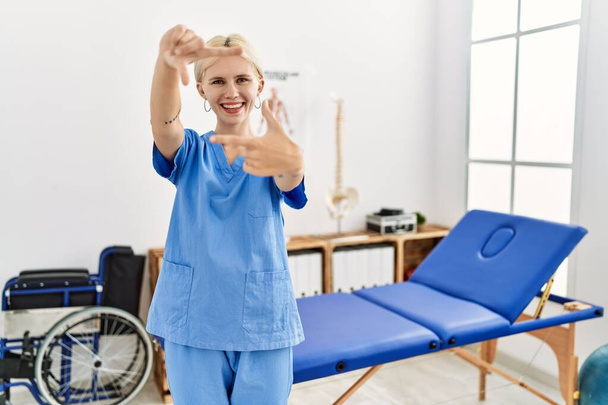 Beautiful caucasian physiotherapist woman working at pain recovery clinic smiling making frame with hands and fingers with happy face. creativity and photography concept.  - Foto, imagen