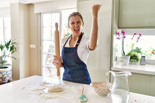 Όμορφη ξανθιά γυναίκα ετοιμάζει ζύμη για πίτσα ενοχλημένη και απογοητευμένη φωνάζοντας με θυμό, φωνάζοντας τρελή από θυμό και σηκωμένη στο χέρι  - Φωτογραφία, εικόνα