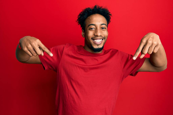 Νεαρός Αφροαμερικάνος με γενειάδα φοράει κόκκινο μπλουζάκι που δείχνει σίγουρος με χαμόγελο στο πρόσωπο, δείχνοντας τον εαυτό του με τα δάχτυλα του περήφανα και χαρούμενα.  - Φωτογραφία, εικόνα