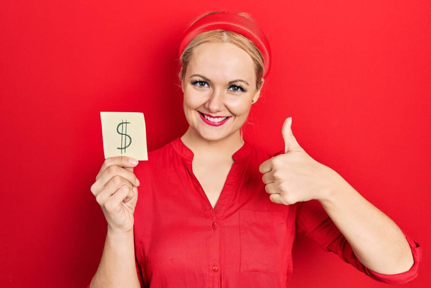 Young blonde woman holding dollar symbol reminder paper smiling happy and positive, thumb up doing excellent and approval sign  - Fotografie, Obrázek