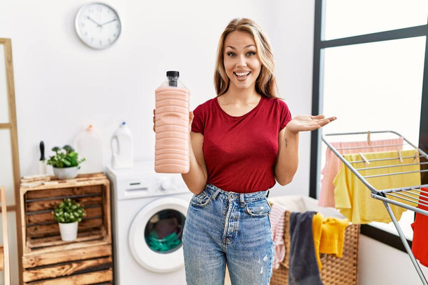 洗濯室で洗剤ボトルを持っている若い白人女性が笑顔で達成を祝い、手を上げて勝者の表情  - 写真・画像