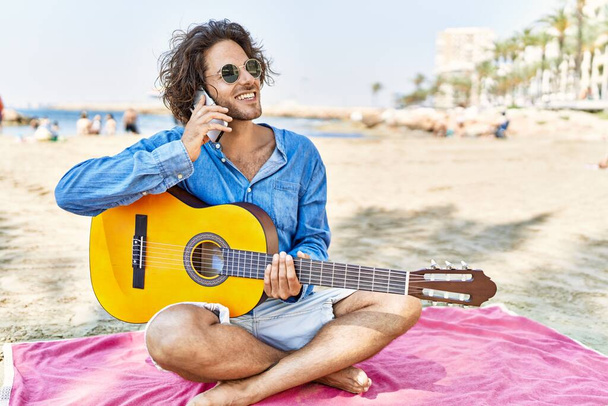 Gitar çalan genç İspanyol adam kumsalda kumların üzerinde oturmuş akıllı telefonla konuşuyor.. - Fotoğraf, Görsel