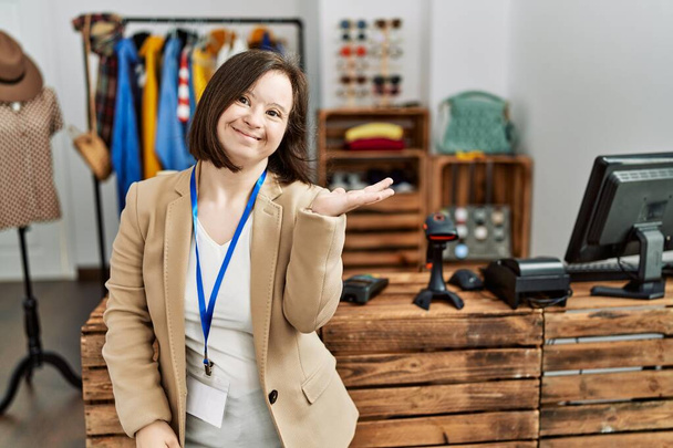 Young down syndrome woman working as manager at retail boutique smiling cheerful presenting and pointing with palm of hand looking at the camera.  - Photo, Image