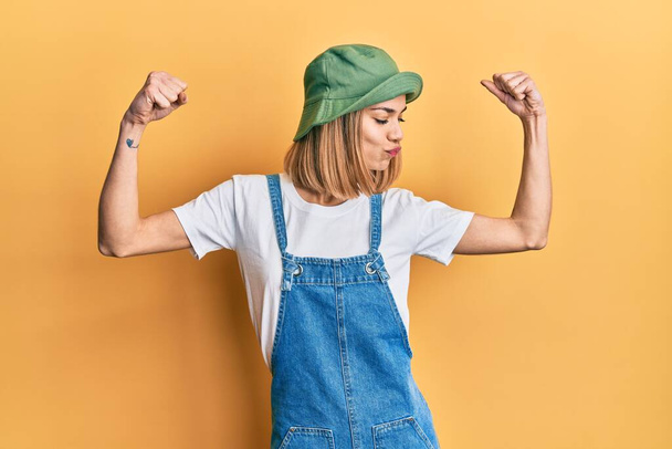 Молодая кавказская блондинка в джинсовом комбинезоне и шляпе в стиле 90-х показывает мышцы рук, улыбающиеся с гордостью. фитнес концепция.  - Фото, изображение