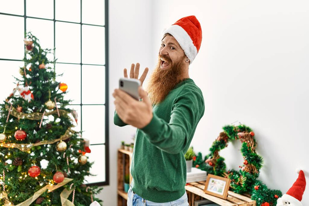 Young redhead man having video call standing by christmas tree at home - Photo, Image