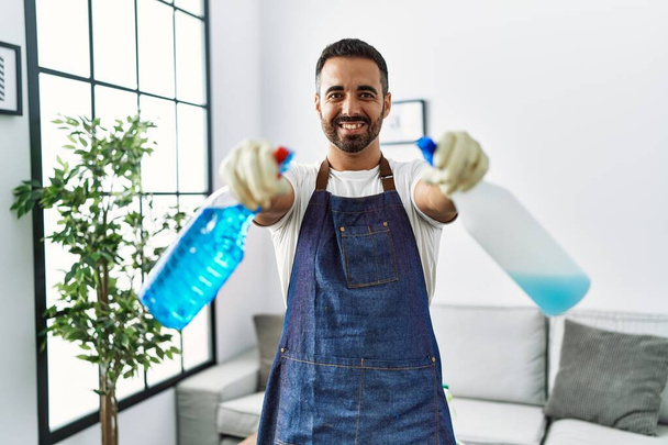 Joven hombre hispano sonriendo confiado sosteniendo pulverizador en casa - Foto, imagen