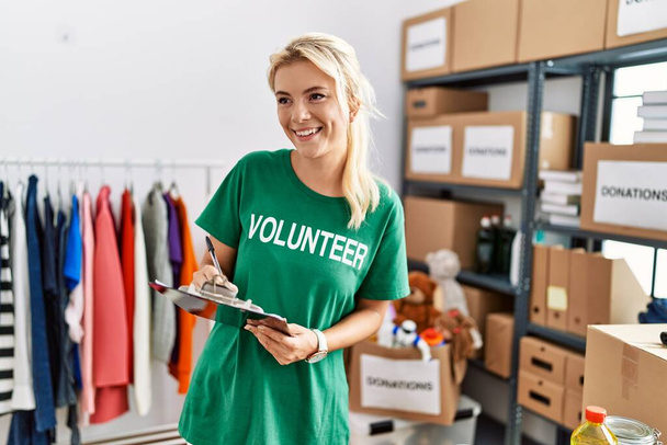 Young blonde volunteer girl smiling happy working at charity center. - 写真・画像