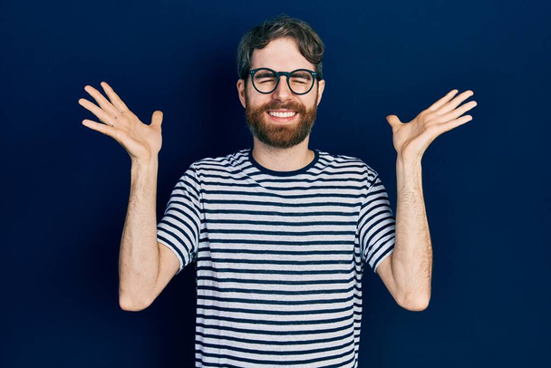 Hombre caucásico con barba vistiendo camiseta a rayas y gafas celebrando loco y loco por el éxito con los brazos levantados y los ojos cerrados gritando emocionado. concepto ganador  - Foto, imagen