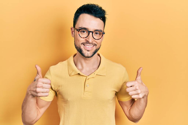 Young hispanic man wearing casual clothes and glasses success sign doing positive gesture with hand, thumbs up smiling and happy. cheerful expression and winner gesture.  - Photo, image