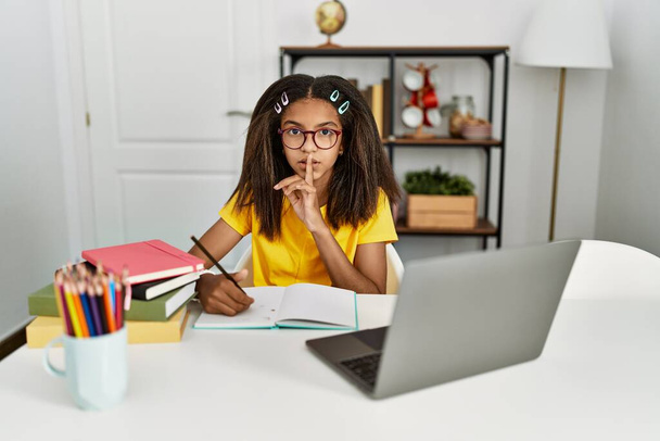 Jeune fille afro-américaine faisant ses devoirs à la maison demandant de se taire avec le doigt sur les lèvres. silence et concept secret.  - Photo, image