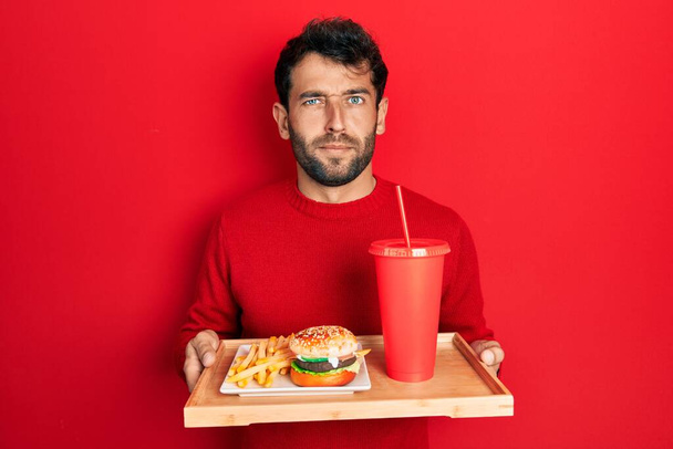 Handsome man with beard eating a tasty classic burger with fries and soda skeptic and nervous, frowning upset because of problem. negative person.  - Фото, зображення