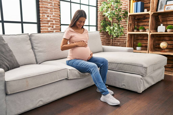 Młoda latynoska kobieta w ciąży uśmiecha się pewnie siedząc na kanapie w domu - Zdjęcie, obraz