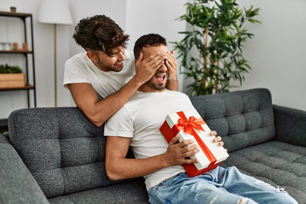 Dva hispánští muži pár překvapení s dárkem a ruce na očích doma - Fotografie, Obrázek