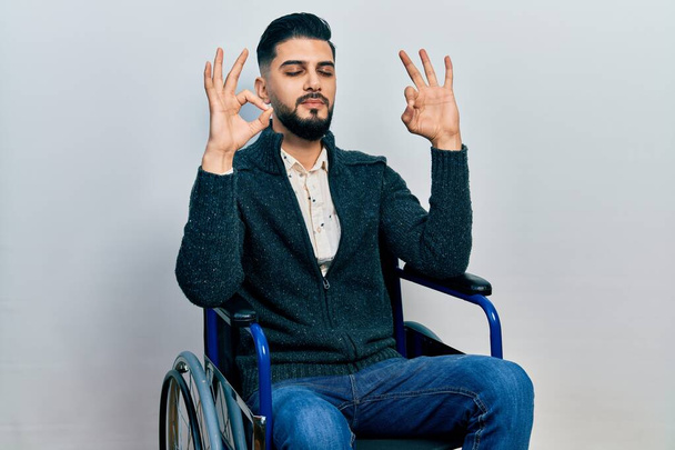 Handsome man with beard sitting on wheelchair relax and smiling with eyes closed doing meditation gesture with fingers. yoga concept.  - Foto, Imagen