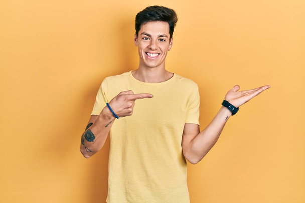 Fiatal spanyol férfi alkalmi sárga pólót visel meghökkent és mosolyog a kamera, miközben bemutatja a kezét, és ujjal mutogatott.  - Fotó, kép