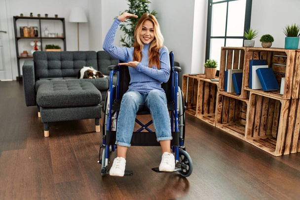 Νεαρή όμορφη γυναίκα κάθεται σε αναπηρική καρέκλα στο σπίτι gesturing με τα χέρια που δείχνουν μεγάλο και μεγάλο μέγεθος σημάδι, μέτρο σύμβολο. Χαμογελώντας κοιτάζοντας την κάμερα. έννοια μέτρησης.  - Φωτογραφία, εικόνα