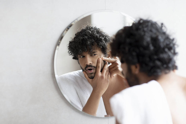 Problémás bőr. Aggódó indiai férfi popping pattanás az arcon, miközben áll a tükör közelében a fürdőszobában - Fotó, kép