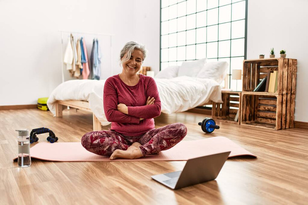 Middle age woman with grey hair training at home looking at exercise video on laptop happy face smiling with crossed arms looking at the camera. positive person.  - Foto, imagen