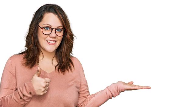 Young plus size woman wearing casual clothes and glasses showing palm hand and doing ok gesture with thumbs up, smiling happy and cheerful  - Foto, imagen