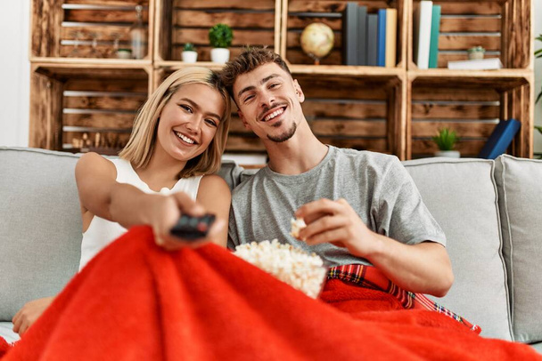 joven caucásico pareja sonriendo feliz viendo película comer palomitas de maíz en casa. - Foto, Imagen