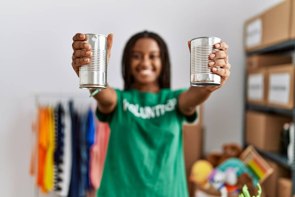 Young african american woman wearing volunteer uniform holding canned food at charity center - Foto, imagen