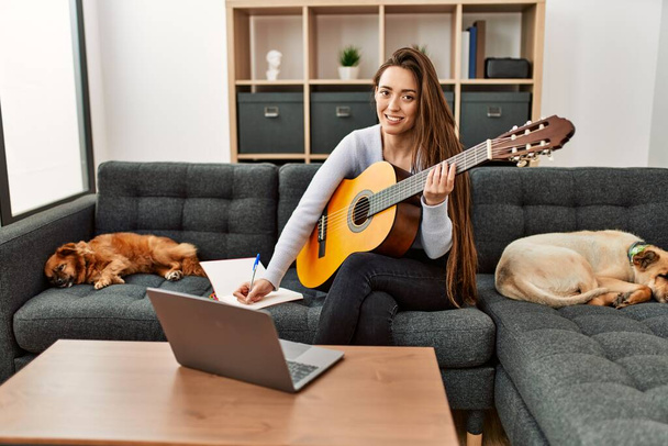 Młoda Latynoska kobieta o online gitara klasy siedzi na kanapie z psami w domu - Zdjęcie, obraz