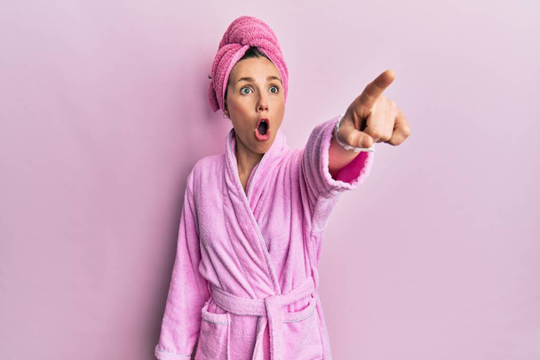 Young blonde woman wearing shower towel cap and bathrobe pointing with finger surprised ahead, open mouth amazed expression, something on the front  - Fotó, kép