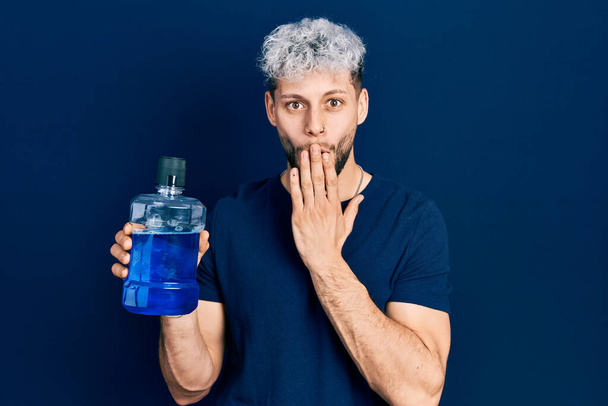 Young hispanic man with modern dyed hair holding mouthwash for fresh breath covering mouth with hand, shocked and afraid for mistake. surprised expression  - Photo, Image