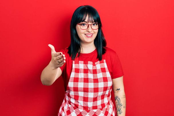 Mujer hispana joven vistiendo delantal de cocinero y gafas sonrientes amistosas ofreciendo apretón de manos como saludo y bienvenida. negocios exitosos.  - Foto, imagen