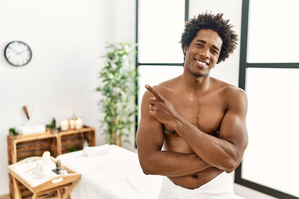 Jovem afro-americano sem camisa usando toalha de pé no centro de beleza com um grande sorriso no rosto, apontando com a mão e o dedo para o lado olhando para a câmera.  - Foto, Imagem