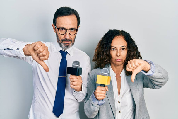 Middle age couple of hispanic woman and man holding reporter microphones with angry face, negative sign showing dislike with thumbs down, rejection concept  - Φωτογραφία, εικόνα