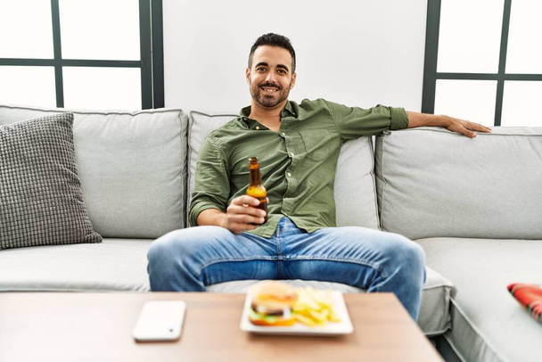 Nuori latino mies syö hampurilainen katsomassa televisiota kotona - Valokuva, kuva