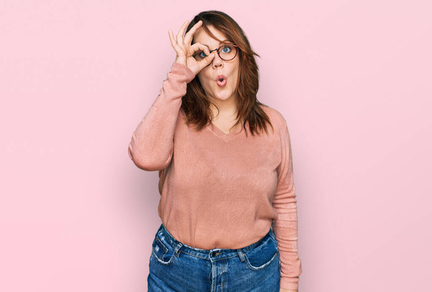 Young plus size woman wearing casual clothes and glasses doing ok gesture shocked with surprised face, eye looking through fingers. unbelieving expression.  - Foto, afbeelding