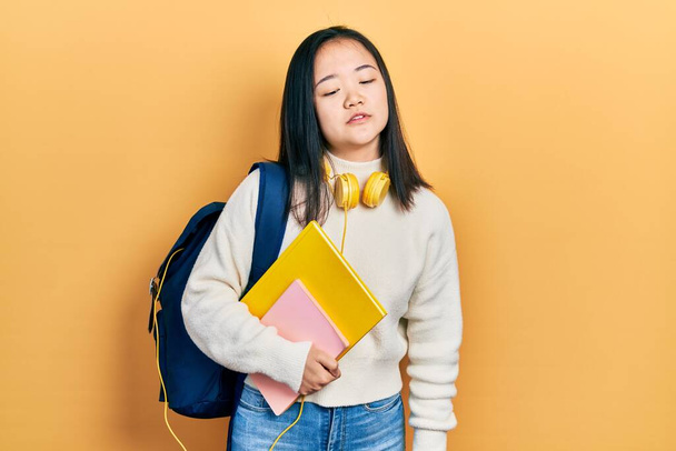 Young chinese girl holding student backpack and books looking sleepy and tired, exhausted for fatigue and hangover, lazy eyes in the morning.  - Zdjęcie, obraz